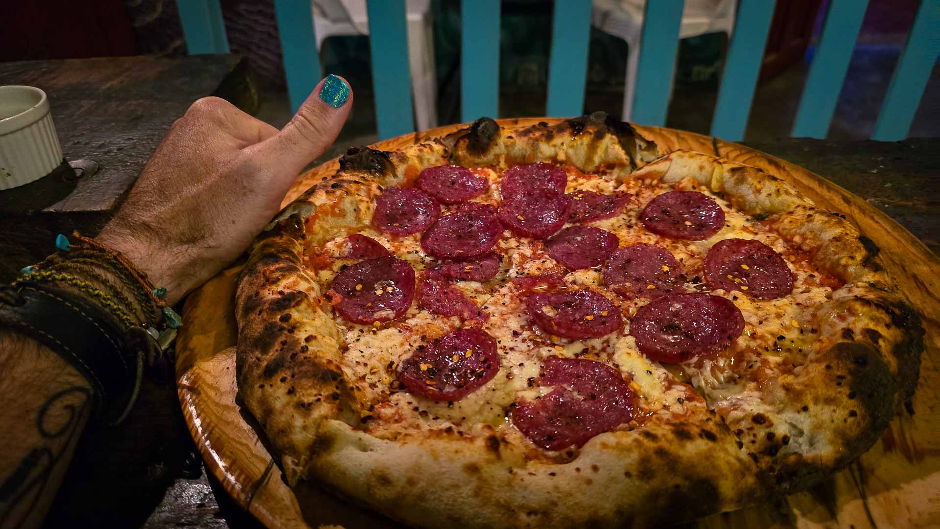 A hand with painted nails and bracelets gives a thumbs up next to a freshly baked pepperoni pizza on a wooden tray, with a blue and white fence in the background.