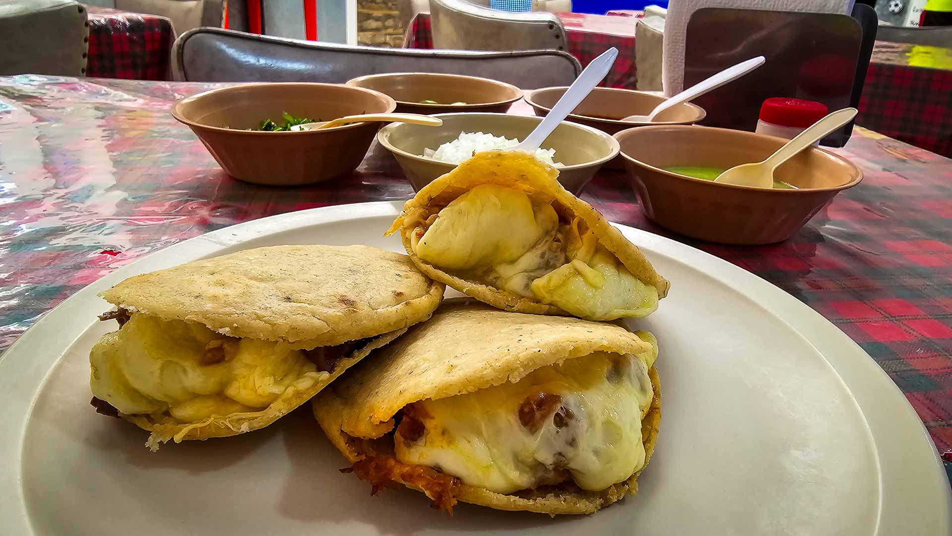 Three cheese-filled gorditas sit on a plate, with bowls of salsa and garnishes in the background on a table covered with a colorful tablecloth.