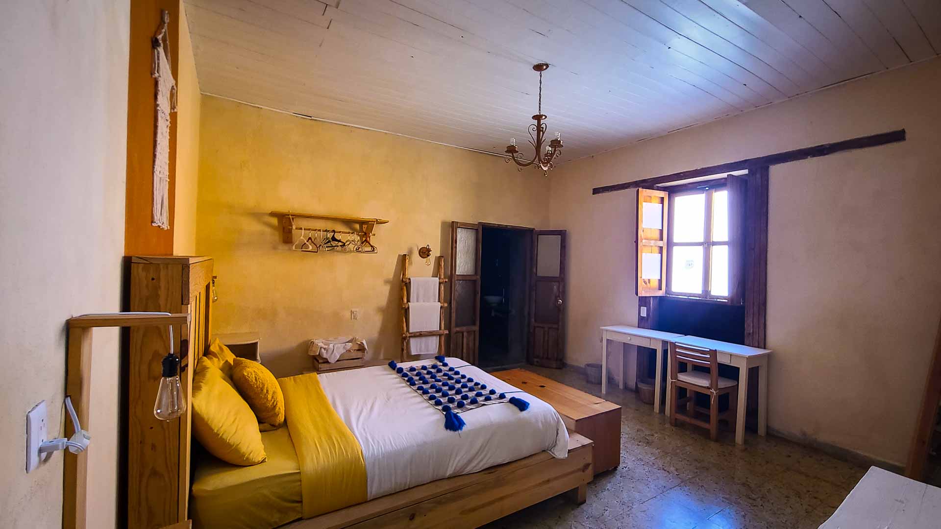 Cozy bedroom with a double bed featuring yellow and white bedding, a wooden headboard, a window with natural light, a desk with two chairs, wall shelves, and a chandelier on the white ceiling.