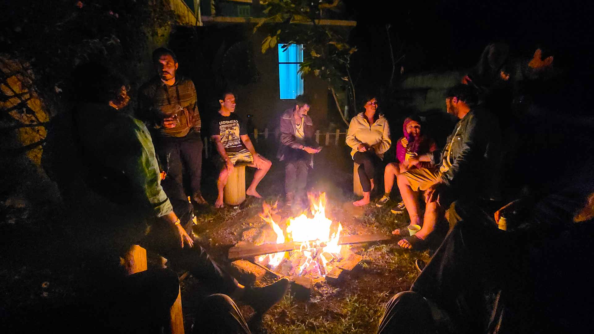 A group of people sit in a circle around a campfire at night, talking and relaxing. The fire illuminates their faces, and a building with a lit blue window is visible in the background.