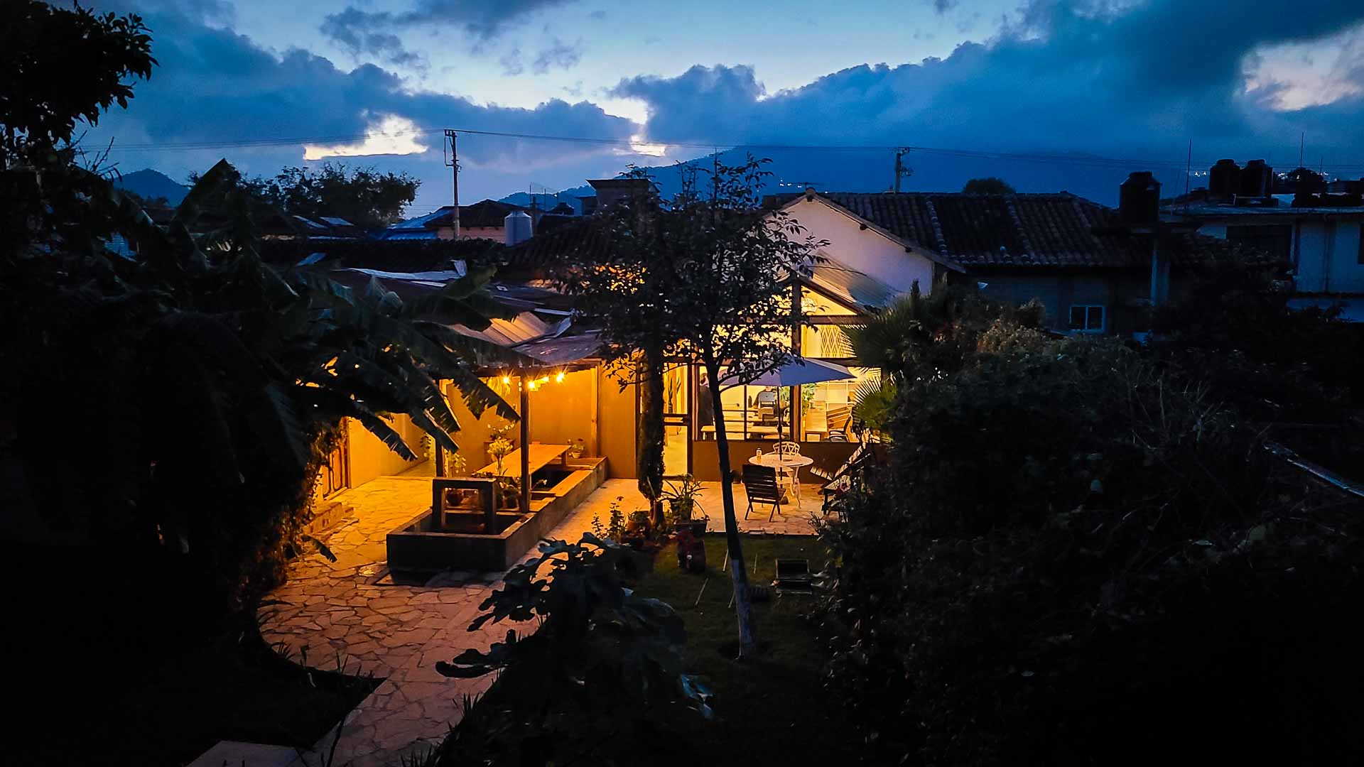 Co404 Coliving house with warm yellow lights glows at dusk, surrounded by lush greenery and trees. The sky is blue with clouds, and a stone path leads to a lit patio area with a table and umbrella.