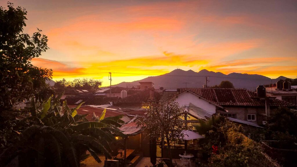Sunset over San Cristobal de las Casas with tiled rooftops, trees, and mountains in the background. The sky is filled with vibrant orange and yellow hues, casting a warm glow across the scene.