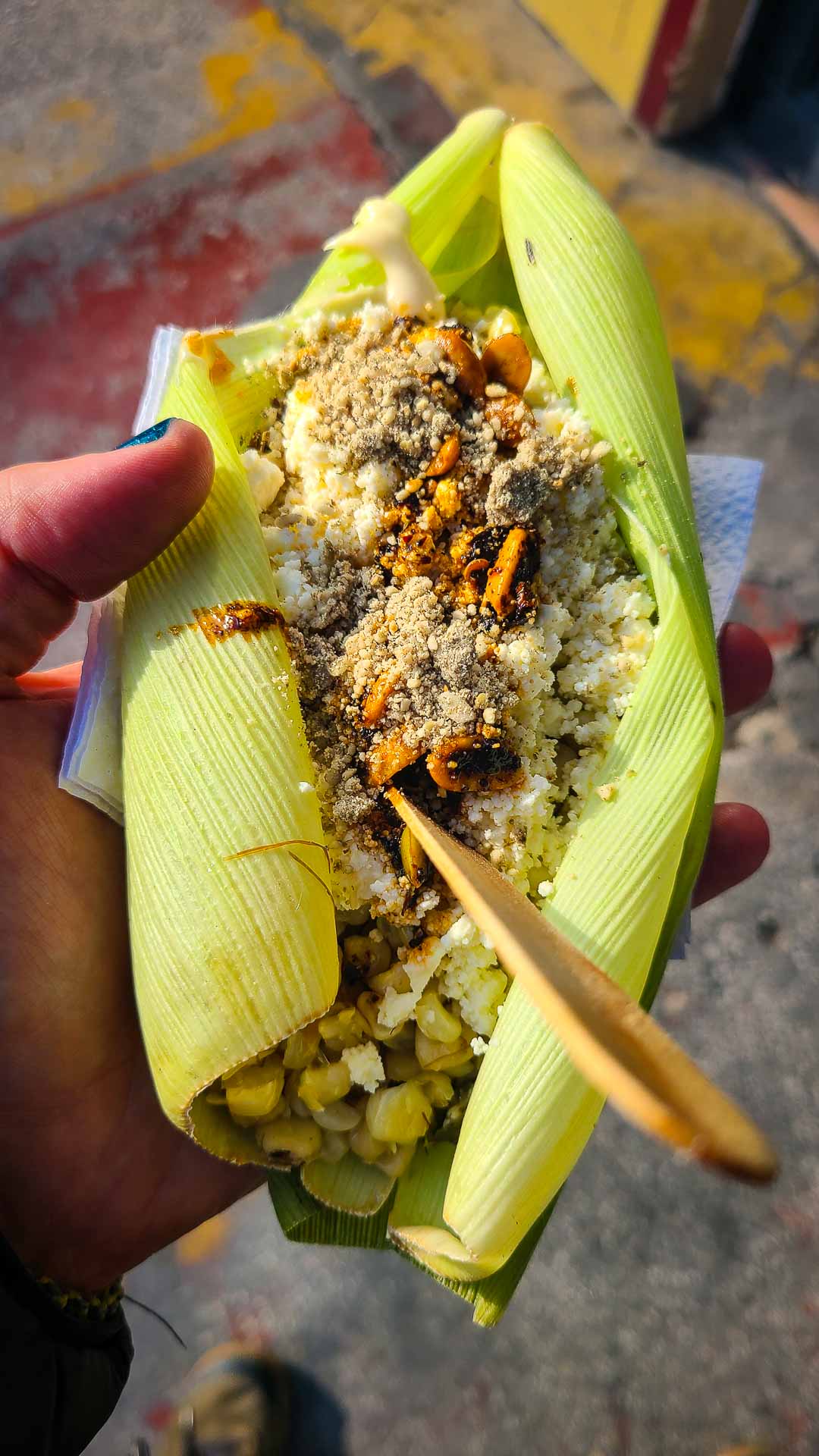A hand holds Mexican street corn (esquites) served in a corn husk, topped with cheese, chili powder, and mayo, with a wooden spoon on top. The street pavement is visible in the background.