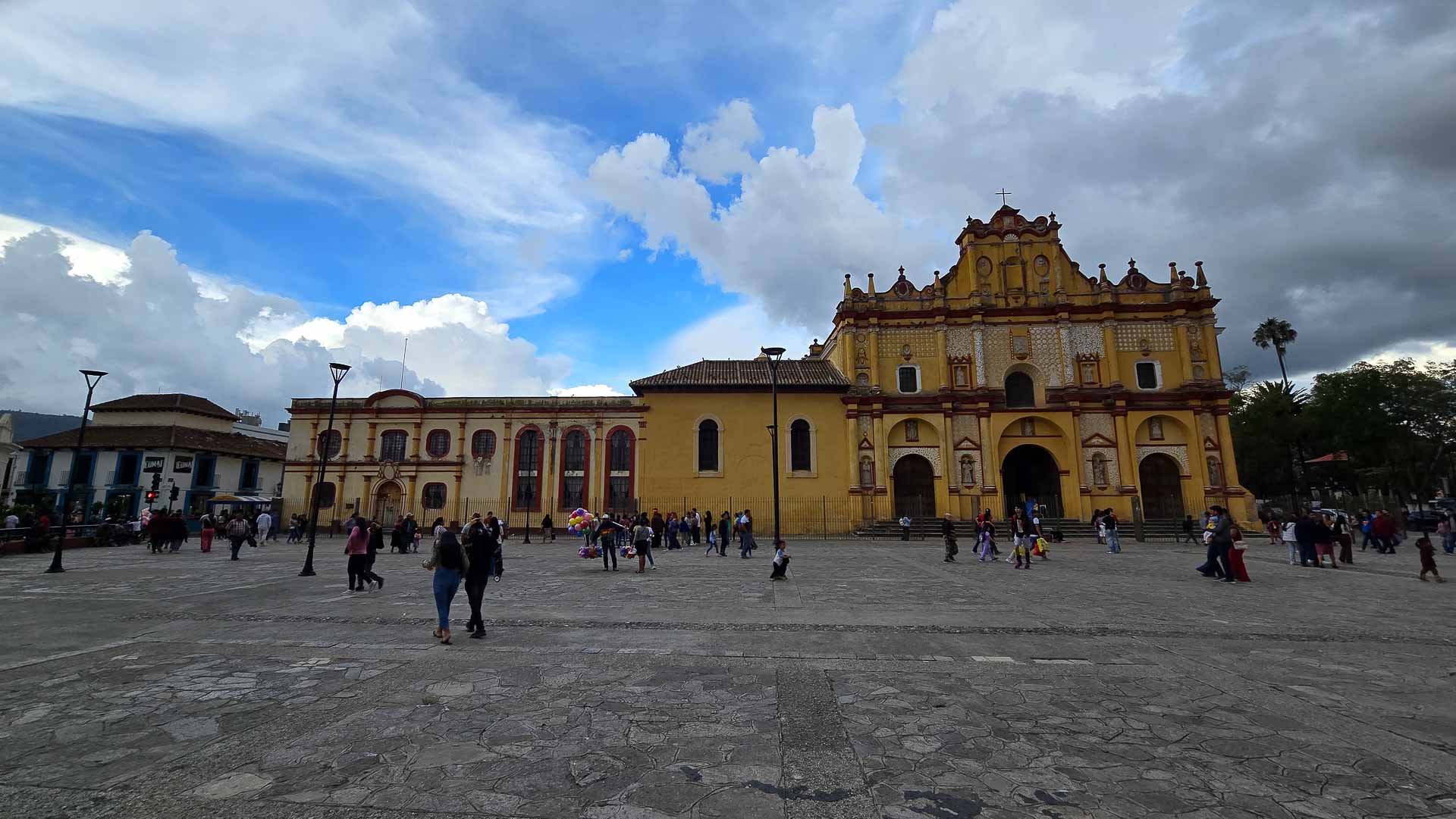 A wide stone plaza with people walking in front of a large yellow colonial-style San Cristobal Cathedral church under a partly cloudy blue sky.