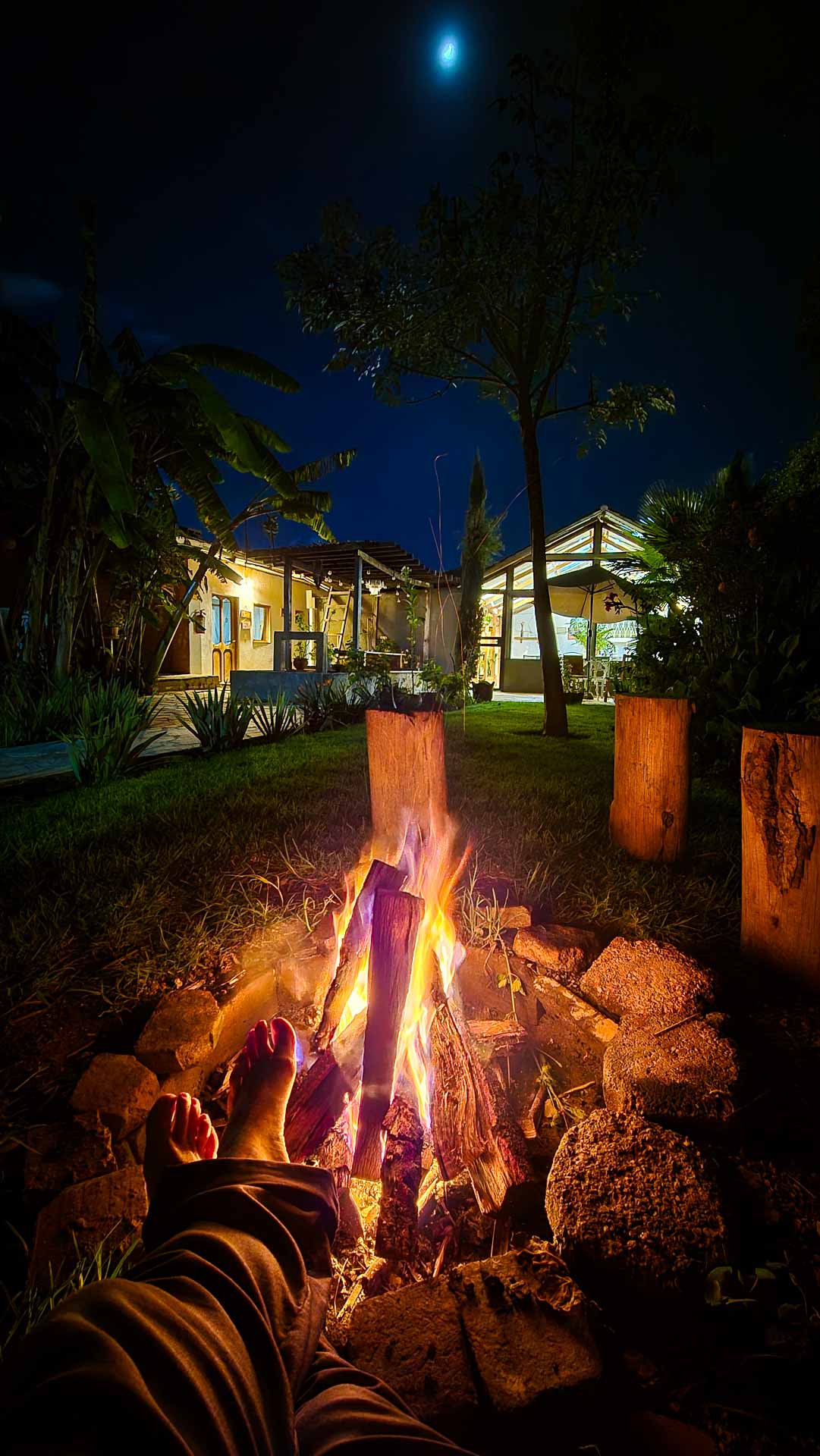 Person relaxing by the Co404 Coliving fire pit at night, with bare feet near the flames, gazing at a well-lit house surrounded by trees and plants under a dark, moonlit sky.