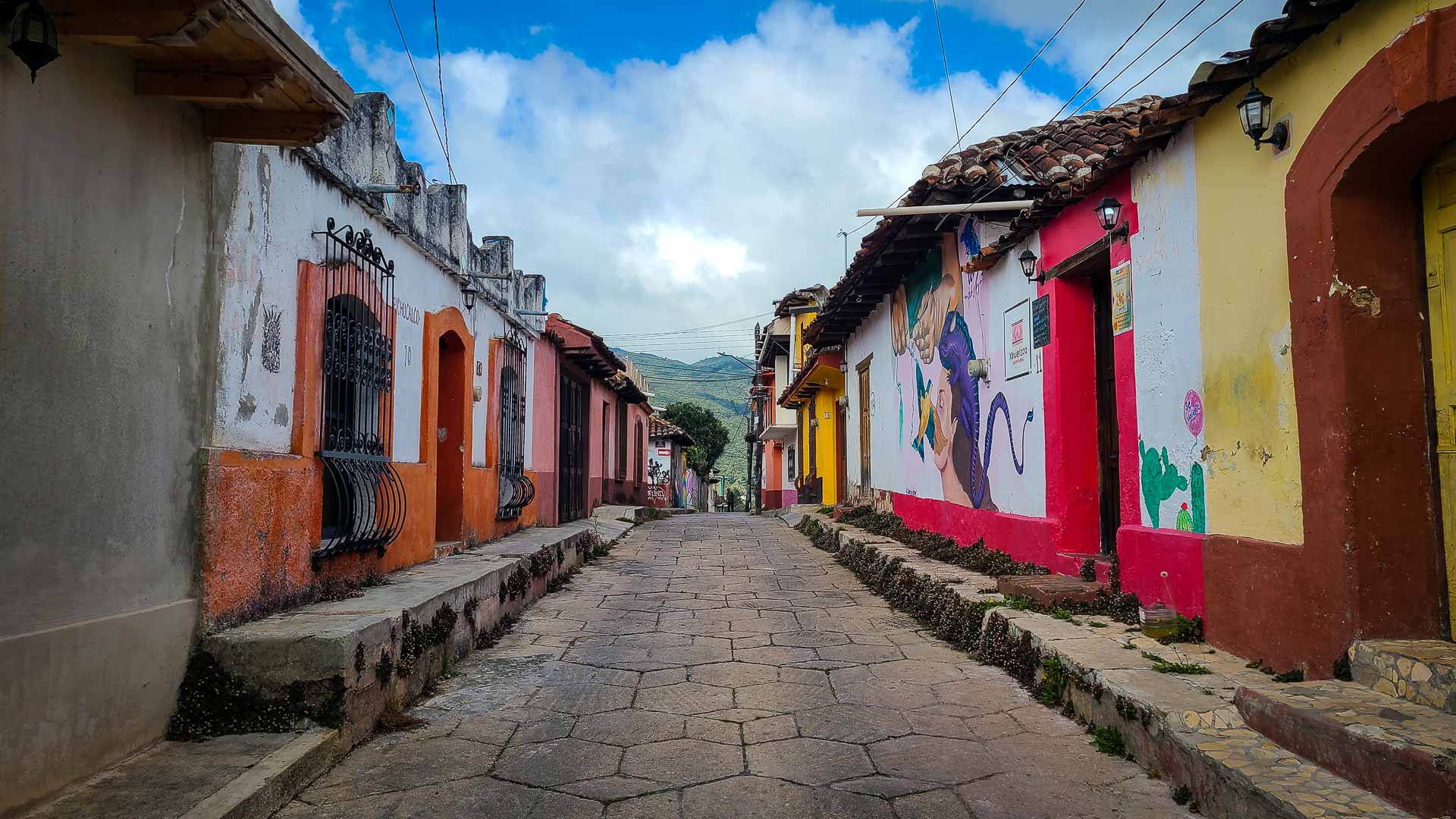A narrow cobblestone street lined with colorful, rustic houses and murals, under a partly cloudy blue sky, in San Cristobal Mexico.