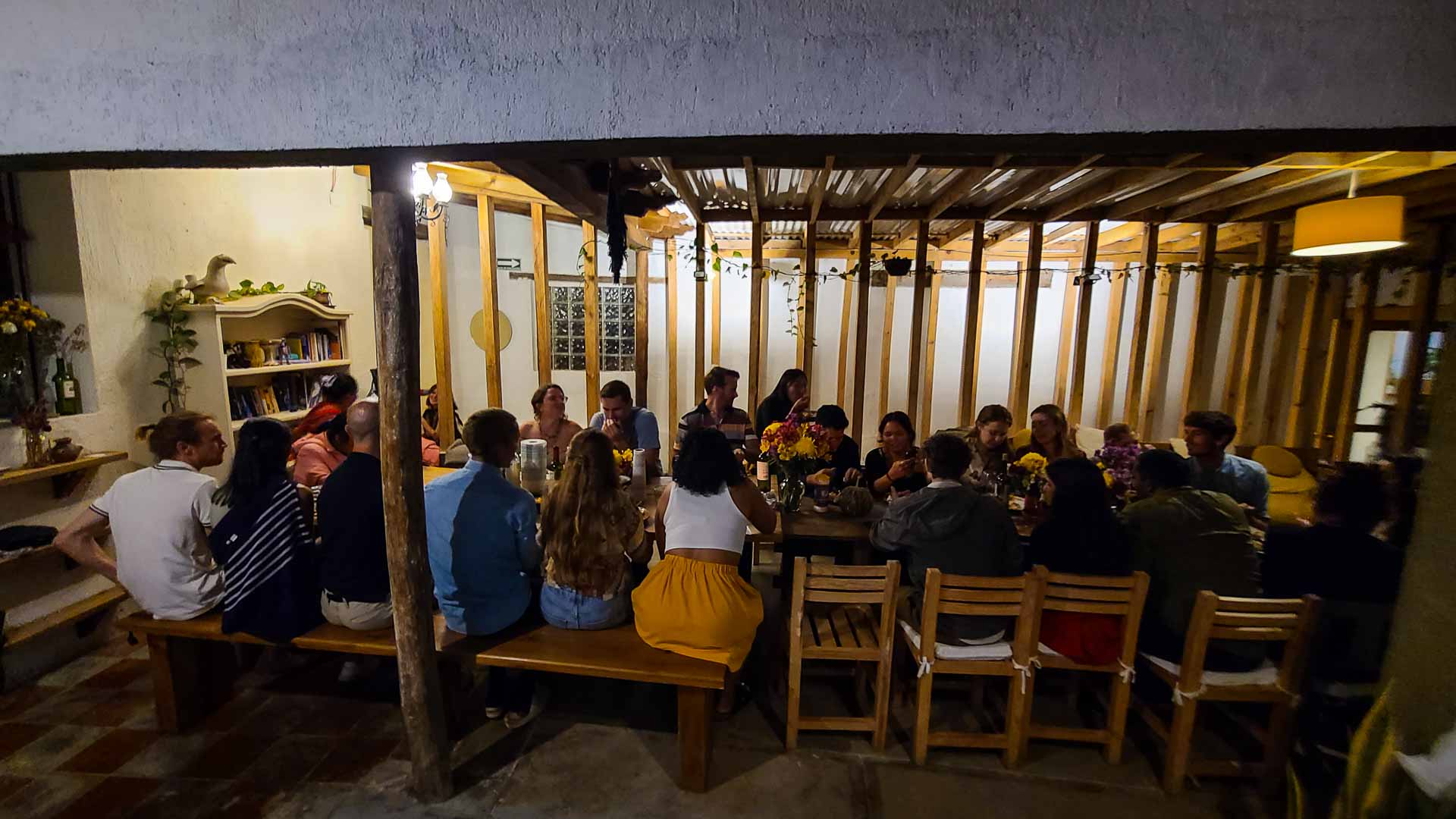 A group of people enjoy Co404 Coliving family dinner around a large dining table in a cozy, warmly lit room with wooden beams. Vases of flowers decorate the table, and bookshelves and plants fill the background.