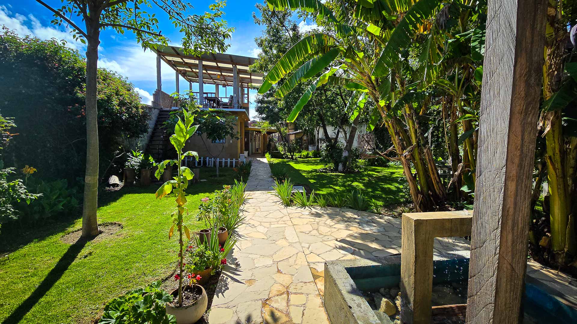 A sunlit garden with lush green grass, tropical plants, and a stone path leads to a two-story Co404 Coliving terrace. Flower pots and a small concrete fountain add to the serene outdoor atmosphere.
